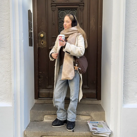 Person standing on a doorstep holding a coffee cup, wearing a coat and scarf.